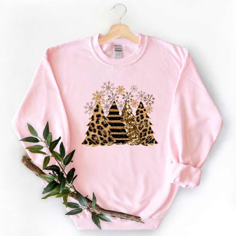 May include: Pink sweatshirt with a graphic of three Christmas trees. The trees are decorated with leopard print, gold glitter, and black and gold stripes. There are gold snowflakes around the trees.