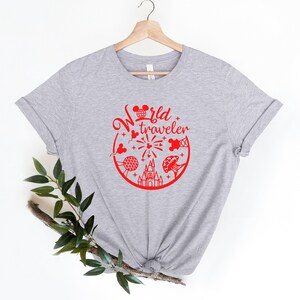 May include: A light gray t-shirt with a red graphic that says "World Traveler." The graphic includes iconic images of a mouse head, a globe, fireworks, a castle, and other travel-related elements. The shirt is displayed on a wooden hanger.