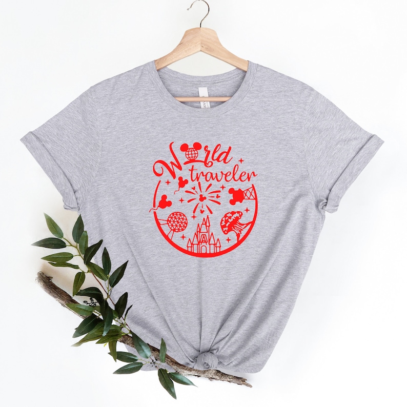 May include: A light gray t-shirt with a red graphic that says "World Traveler." The graphic includes iconic images of a mouse head, a globe, fireworks, a castle, and other travel-related elements. The shirt is displayed on a wooden hanger.