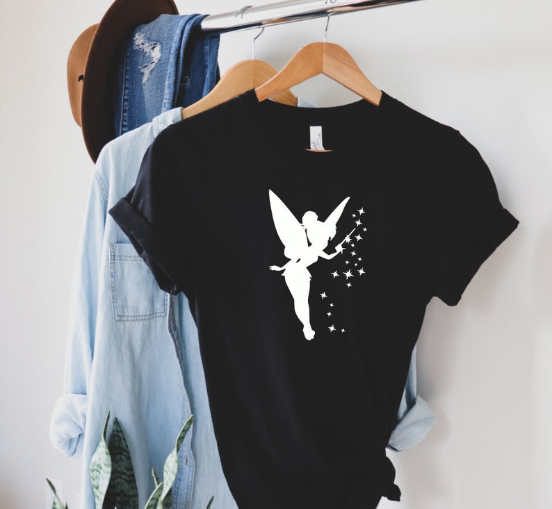 Puede incluir: Camiseta negra con una silueta blanca de un hada con una varita y estrellas.