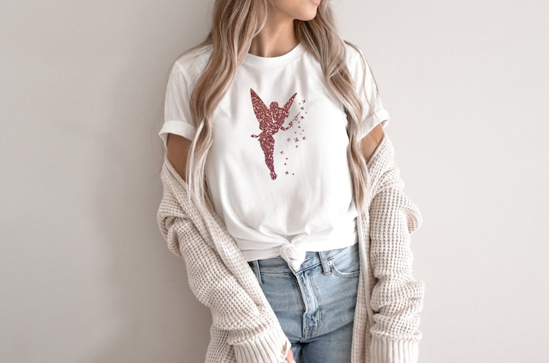 Puede incluir: Una camiseta blanca con un dise&ntilde;o de hada en purpurina rosa y estrellas. El hada sostiene una varita m&aacute;gica y tiene alas. La camiseta est&aacute; anudada a la cintura y la modelo lleva pantalones vaqueros azules y una chaqueta de punto crema.