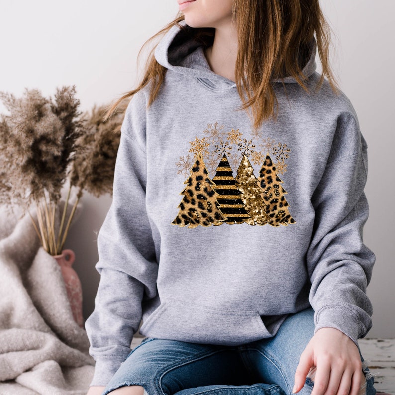 May include: A gray hooded sweatshirt with a design of three Christmas trees. The trees are decorated with leopard print, black and white stripes, and gold glitter. There are gold snowflakes around the trees.