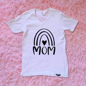 Op de afbeelding: Wit T-shirt met V-hals en een zwart regenboogontwerp met het woord "MOM" in het midden. Een klein zwart hartje bevindt zich in de regenboog.