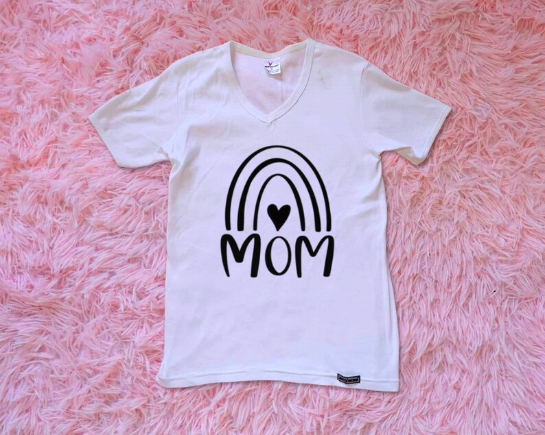 Op de afbeelding: Wit T-shirt met V-hals en een zwart regenboogontwerp met het woord "MOM" in het midden. Een klein zwart hartje bevindt zich in de regenboog.