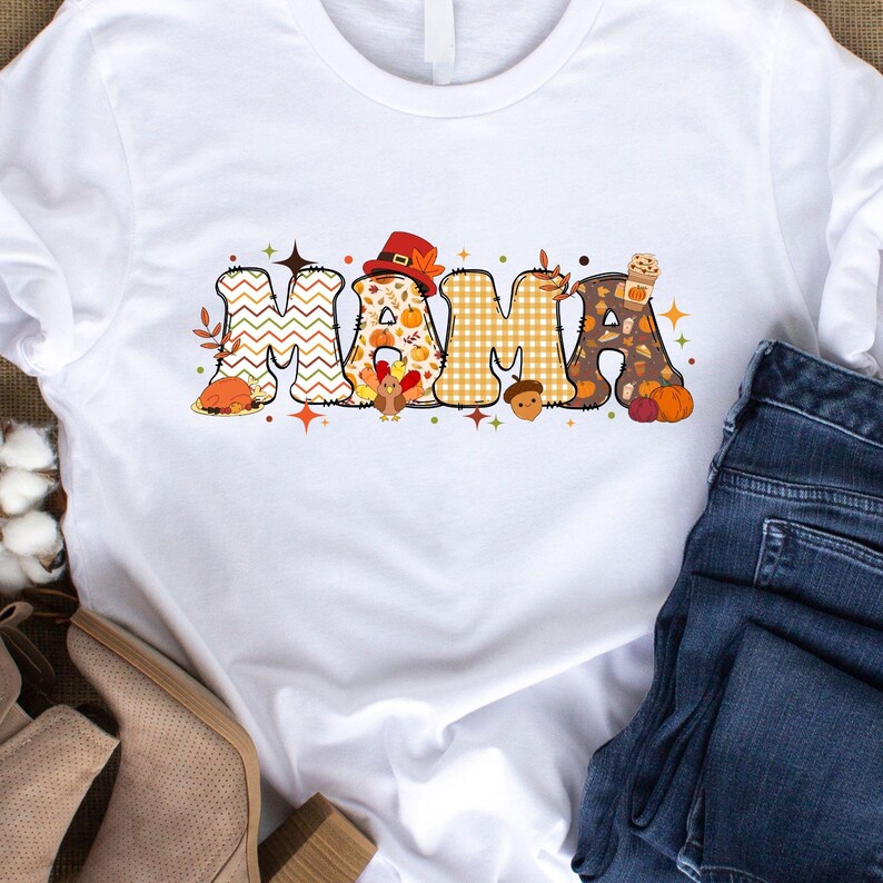 Pu&ograve; includere: Maglietta bianca con la parola "MAMA" in lettere di patchwork colorate con disegni a tema autunnale, tra cui tacchini, zucche e ghiande.