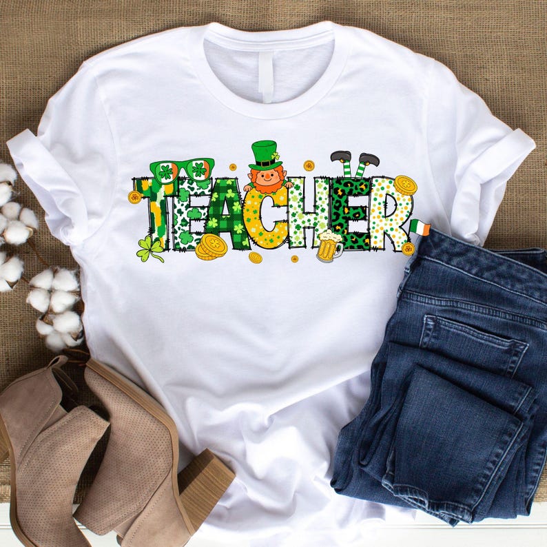 Puede incluir: Camiseta blanca con un gr&aacute;fico verde, amarillo y negro que dice "Teacher" con un duende y otras im&aacute;genes con tem&aacute;tica del D&iacute;a de San Patricio.