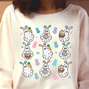 Puede incluir: Sudadera blanca con un patrón repetido de fantasmas de color pastel que sostienen cestas de huevos de Pascua. Los fantasmas tienen orejas de conejo y están decorados con lunares de colores.