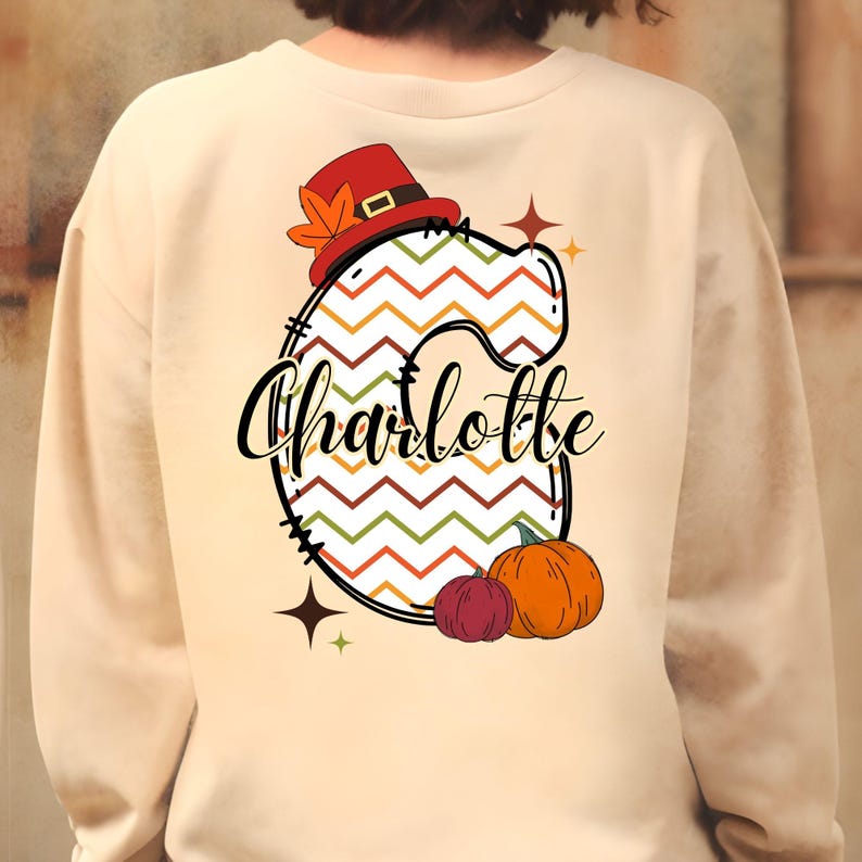 Pu&ograve; includere: Una felpa beige con una grande lettera "C" colorata a motivo chevron, un cappello da pellegrino e foglie autunnali. Il nome "Charlotte" &egrave; scritto in un font corsivo nero sotto la lettera. Due piccole zucche si trovano sotto la lettera.