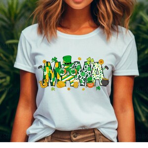Puede incluir: Camiseta blanca con un gr&aacute;fico verde, amarillo y dorado que dice "MAMA" con tr&eacute;boles, duendes y otras im&aacute;genes con tem&aacute;tica del D&iacute;a de San Patricio.