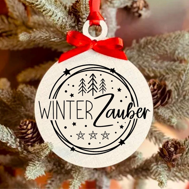 May include: White round Christmas ornament with a black and white design. The design features a circle with three stylized pine trees, stars, and the text "Winter Zauber". The ornament is hanging from a red ribbon.