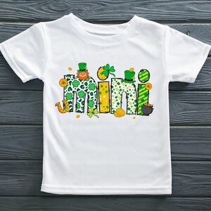 Puede incluir: Camiseta blanca con un gr&aacute;fico verde, amarillo y dorado de la palabra "mini" con tr&eacute;boles, un duende y monedas de oro.