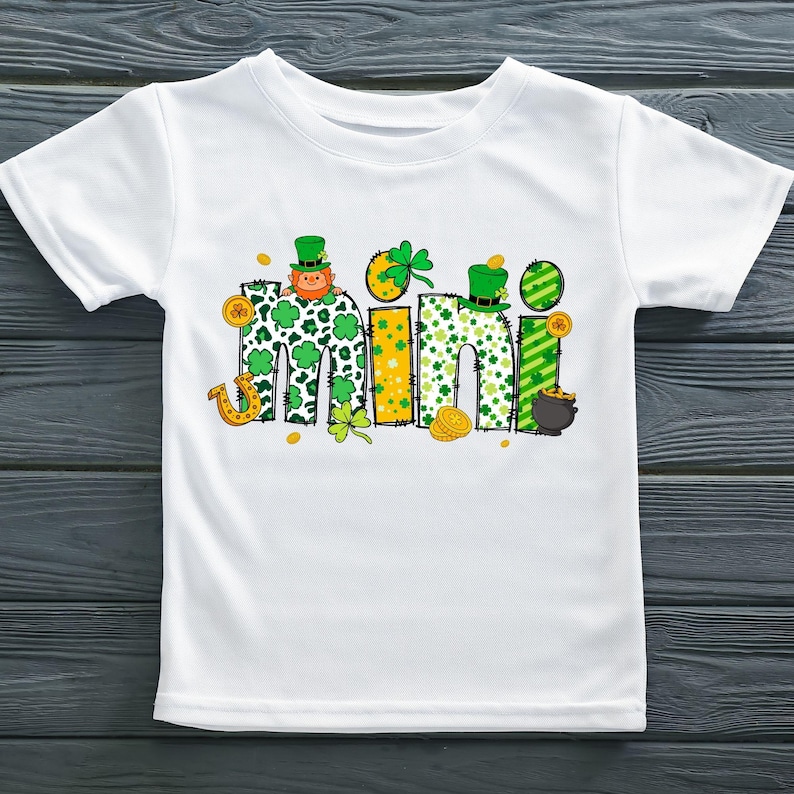 Puede incluir: Camiseta blanca con un gr&aacute;fico verde, amarillo y dorado de la palabra "mini" con tr&eacute;boles, un duende y monedas de oro.