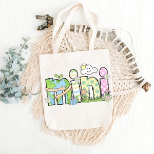 May include: A white canvas tote bag with a colorful design featuring the word "mini" with a cartoon earth hugging the "m". The design also includes a sun, a cloud, flowers, and a watering can.