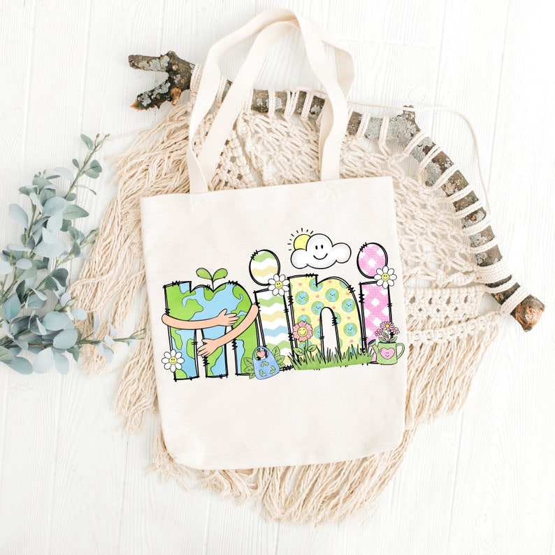 May include: A white canvas tote bag with a colorful design featuring the word "mini" with a cartoon earth hugging the "m". The design also includes a sun, a cloud, flowers, and a watering can.