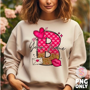 K&ouml;nnte beinhalten: Ein cremefarbener Pullover mit einem pinkfarbenen und goldenen Grafikdesign, das einen Buchstaben B aus Herzen und Pailletten zeigt. Die Grafik enth&auml;lt auch einen kleinen Umschlag mit einem Herzen und dem Text "Your Name".