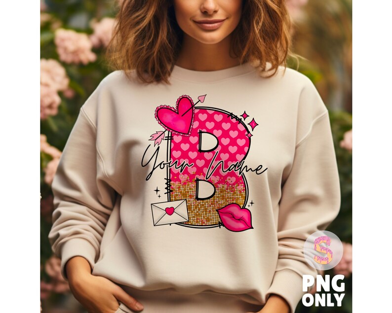 K&ouml;nnte beinhalten: Ein cremefarbener Pullover mit einem pinkfarbenen und goldenen Grafikdesign, das einen Buchstaben B aus Herzen und Pailletten zeigt. Die Grafik enth&auml;lt auch einen kleinen Umschlag mit einem Herzen und dem Text "Your Name".