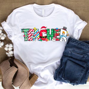 Peut inclure: T-shirt blanc avec un graphisme de No&euml;l "TEACHER". Les lettres sont d&eacute;cor&eacute;es de cannes en sucre, de boules, de flocons de neige et du P&egrave;re No&euml;l.