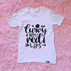 May include: White V-neck t-shirt with the text "Curvy Hips Red Lips" in black script. The design includes a lipstick, a kiss mark, and an arrow. The shirt is displayed on a pink, fluffy surface.