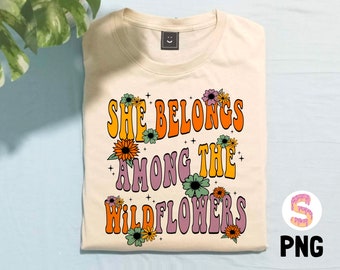 May include: A beige t-shirt with the text "She Belongs Among The Wildflowers" in a retro font with colorful flowers surrounding the text.