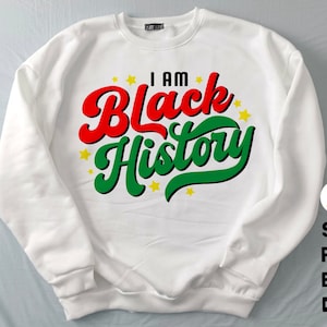 Puede incluir: Sudadera blanca con el texto "I AM Black History" en rojo y verde, con estrellas amarillas. La sudadera es de cuello redondo.
