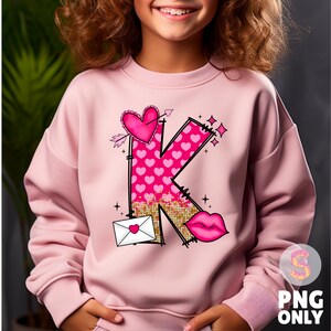 K&ouml;nnte beinhalten: Rosafarbener Pullover mit einem Grafikdruck eines Buchstabens K aus rosa Herzen und Glitzer. Der Buchstabe ist von Herzen, Pfeilen und einem Lippenstift-Kuss umgeben.
