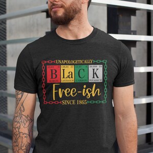 Pu&ograve; includere: Una maglietta nera con un design grafico che presenta la parola "Black" scritta usando simboli di elementi chimici. La grafica include anche il testo "Free-ish since 1865" e un design a catena.