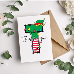Peut inclure: Une carte blanche avec une illustration d'un lutin de No&euml;l vert et rouge. Le lutin porte un chapeau et a une chaussette verte avec des rayures rouges et blanches. La carte dit "Merci" en &eacute;criture cursive noire.