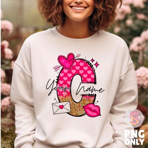 K&ouml;nnte beinhalten: Wei&szlig;er Pullover mit einem pinkfarbenen und goldenen Buchstaben C-Design. Der Buchstabe besteht aus Herzen und Glitzer. Der Text "Your Name" ist in schwarzer Kursivschrift geschrieben.