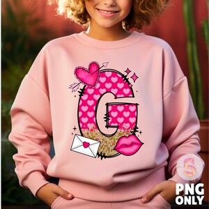 K&ouml;nnte beinhalten: Rosafarbener Pullover mit einem Grafikdruck eines Buchstabens G aus rosa Herzen, einem Herz mit einem Pfeil, einem goldenen Glitzerhintergrund, einer roten Lippe und einem wei&szlig;en Umschlag mit einem rosa Herz. Der Text "G" ist in Schwarz.
