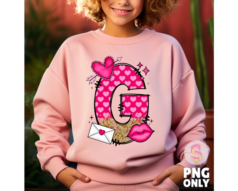 K&ouml;nnte beinhalten: Rosafarbener Pullover mit einem Grafikdruck eines Buchstabens G aus rosa Herzen, einem Herz mit einem Pfeil, einem goldenen Glitzerhintergrund, einer roten Lippe und einem wei&szlig;en Umschlag mit einem rosa Herz. Der Text "G" ist in Schwarz.