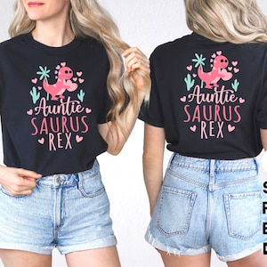 May include: Black t-shirt featuring a pink dinosaur graphic and the text "Auntie Saurus Rex" in white. The dinosaur is holding a pink heart. The shirt is being worn by a person with long blonde hair.