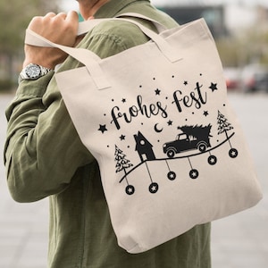 May include: A white canvas tote bag with black handles and the text "frohes fest" in a stylized font. The bag features a black silhouette of a truck carrying a Christmas tree, a house, and stars.