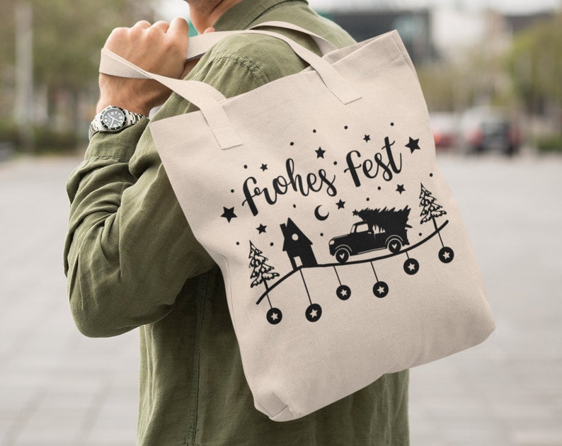 May include: A white canvas tote bag with black handles and the text "frohes fest" in a stylized font. The bag features a black silhouette of a truck carrying a Christmas tree, a house, and stars.