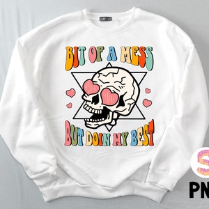 Op de afbeelding: Witte trui met een kleurrijke afbeelding van een schedel met harten als ogen en de tekst "Bit of a mess but doin my best".