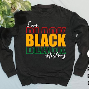 Puede incluir: Sudadera gris oscuro con las palabras "I am BLACK History" en rojo, naranja, verde y blanco. El diseño está en capas, con cada palabra ligeramente desplazada. La sudadera tiene cuello redondo y mangas largas.