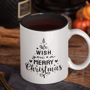 May include: White ceramic coffee mug with a black rim and handle. The mug has a black design of a stylized Christmas tree with the text "We wish you a Merry Christmas".