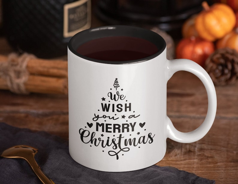 May include: White ceramic coffee mug with a black rim and handle. The mug has a black design of a stylized Christmas tree with the text "We wish you a Merry Christmas".