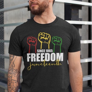 Pu&ograve; includere: Maglietta nera con una grafica di tre pugni alzati in rosso, giallo e verde. Il testo "Since 1865 Freedom Juneteenth" &egrave; stampato sotto i pugni.