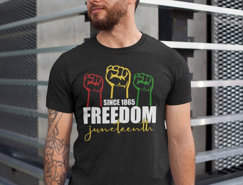 Pu&ograve; includere: Maglietta nera con una grafica di tre pugni alzati in rosso, giallo e verde. Il testo "Since 1865 Freedom Juneteenth" &egrave; stampato sotto i pugni.