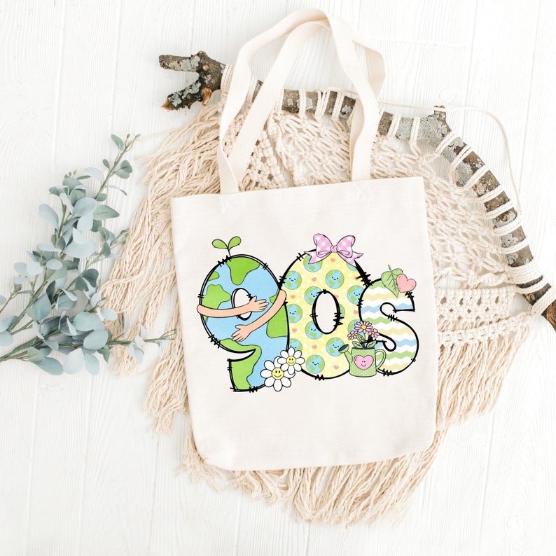 May include: A white tote bag with a design of the numbers 90s, each number is shaped like the Earth with a green outline and a blue and green globe inside. The numbers are decorated with flowers, a watering can, and a bow.