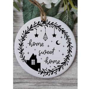May include: White ceramic ornament with a black silhouette of a house, stars, a crescent moon, and a diamond hanging from a wreath. The text "home sweet home" is written in a cursive font.