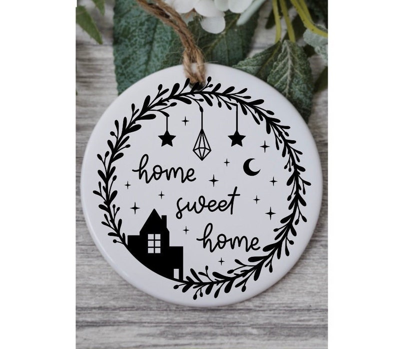 May include: White ceramic ornament with a black silhouette of a house, stars, a crescent moon, and a diamond hanging from a wreath. The text "home sweet home" is written in a cursive font.