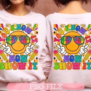 Peut inclure: Un sweat-shirt rose avec un motif graphique coloré représentant un visage souriant avec des lunettes de soleil et le texte "You Know It Test Day Now Show It" sur un fond à carreaux.