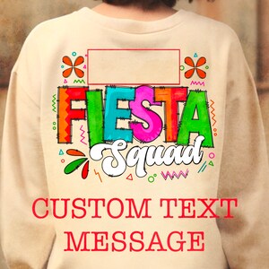 May include: A tan sweatshirt with a colorful "Fiesta Squad" graphic and a blank space for custom text. The graphic features a colorful flower design and a border of zigzags and dots.