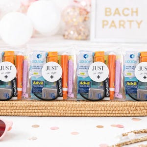 May include: Four clear plastic bags filled with small items, arranged on a woven tray. Each bag has a round sticker that says "JUST in case." Contents include orange and pink tubes, small packets, and a gray item labeled "Advil."