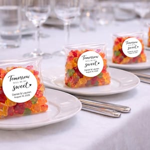 May include: Clear plastic bags filled with multicolored gummy bears, each featuring a circular white sticker that reads Tomorrow WILL BE SO sweet, Daniel & Lauren, August 18, 2026, arranged on white plates.