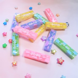 May include: A collection of colorful rectangular erasers in various shades of pink, yellow, green, and blue. Each eraser is wrapped in clear plastic and features text in both English and another language. Decorative star-shaped origami is scattered around the erasers.