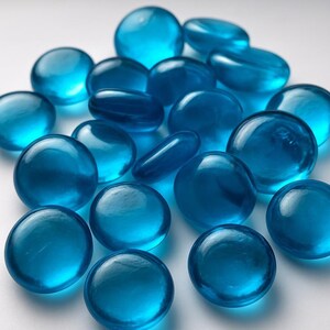 May include: A collection of clear, rounded, and oval-shaped glass beads in various sizes. The beads are a vibrant shade of blue, with a glossy, reflective surface. The arrangement is scattered, creating a visually appealing display.