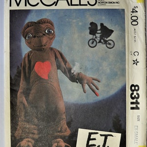 May include: A McCall's sewing pattern envelope for an E.T. the Extra-Terrestrial costume, size EX-SMALL, featuring a brown creature with a red heart on its chest and a glowing red fingertip, set against a large blue moon and a silhouette graphic.