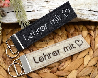 May include: Two rectangular keychains with metal hardware. One is black with white embroidered text and a heart, the other is beige with brown text and a heart. The text reads "Lehrer mit".